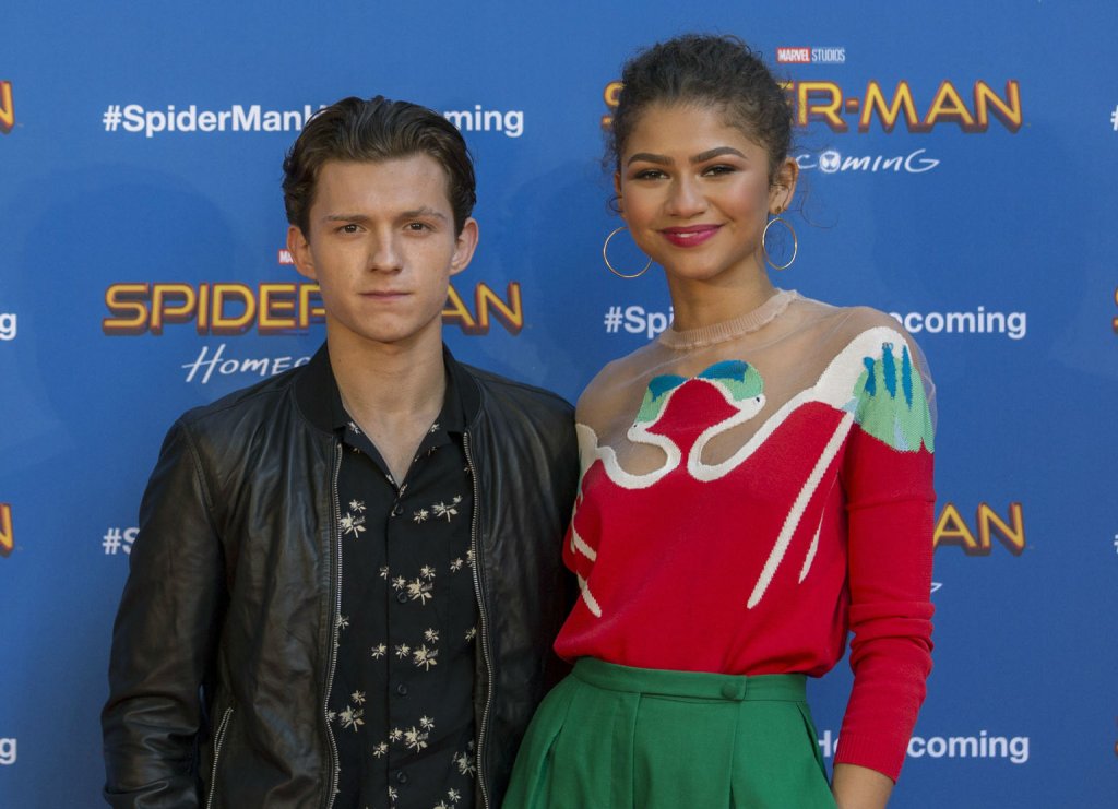 Tom Holland e Zendaya em divulgação de Homem-Aranha; ele está vestindo uma jaqueta de couro preta com camisa da mesma cor com detalhes de flores brancas e Zendaya está com um look verde e vermelho; os dois estão abraçados e ela sorri levemente enquanto ele tem a expressão mais séria