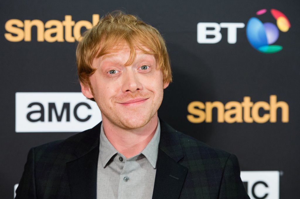 Rupert Grint em carpet de premiere sorrindo levemente para a câmera e arregalando os olhos; ele usa uma camisa cinza com blazer escuro