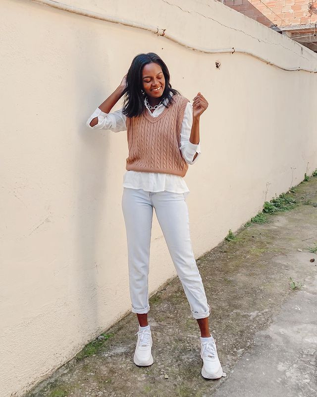 look-inverno-sobreposicao-colete-trico-dayaneassiis Garota usando colete de tricô marrom por cima de camisa branca, com calça jeans clara e tênis branco. Ela está sorrindo, com uma das mãos atrás do cabelo e o outro braço dobrado para cima.