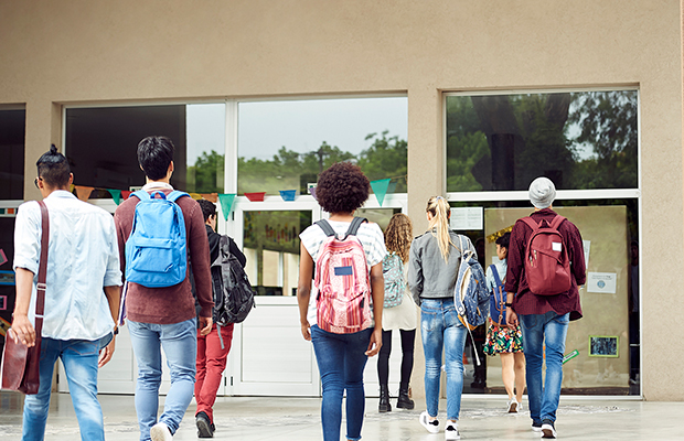 A imagem mostra várias pessoas, com mochilas nas costas, viradas de costas e caminhando em direção a entrada do que parece ser uma escola.