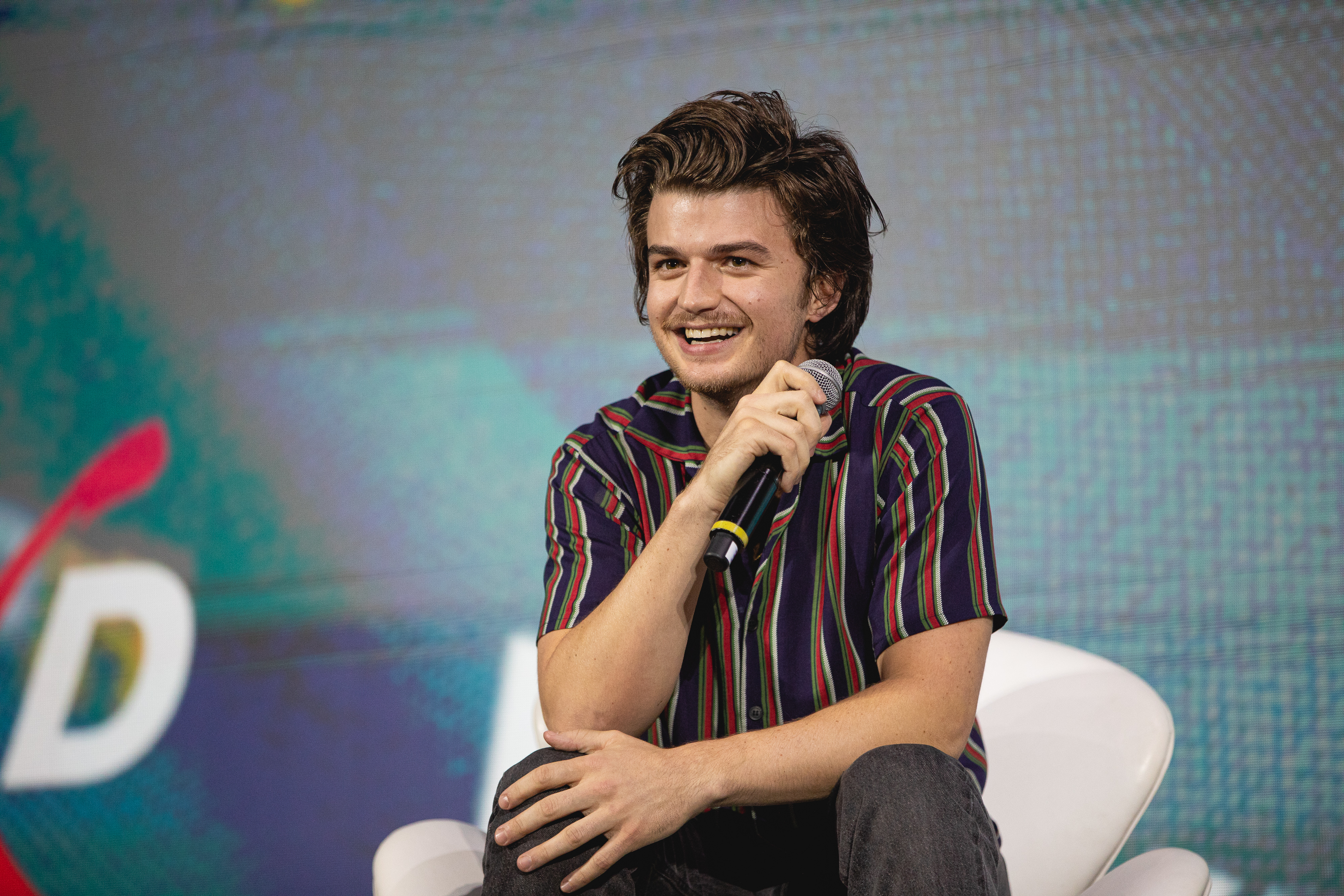 Joe Keery sentado em cadeira branca no palco do painel de Free Guy, na CCXP 2019, em São Paulo; o ator segura o microfone com uma das mãos enquanto a outra repousa em seu joelho, ele sorri observando o público e usa uma camisa listrada em tons de azul escuro, vermelho e verde