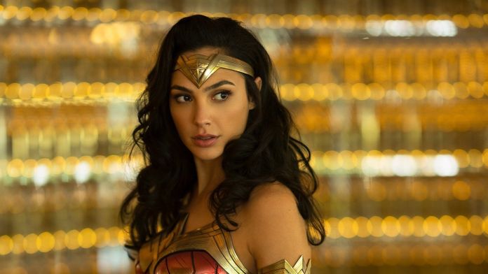 Foto da atriz Gal Gadot como a super-heroína Mulher-Maravilha. Nela, ela aparece com um acessório dourado na testa, roupa dourada e vermelha e fundo de luzes amarelas.