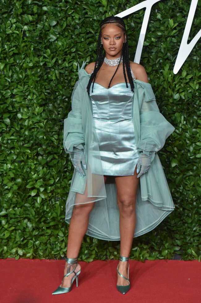 Rihanna posando com trança, vestido azul e casado ca mesma cor. Em tapete vermelho com fundo verde. Sua expressão é séria.