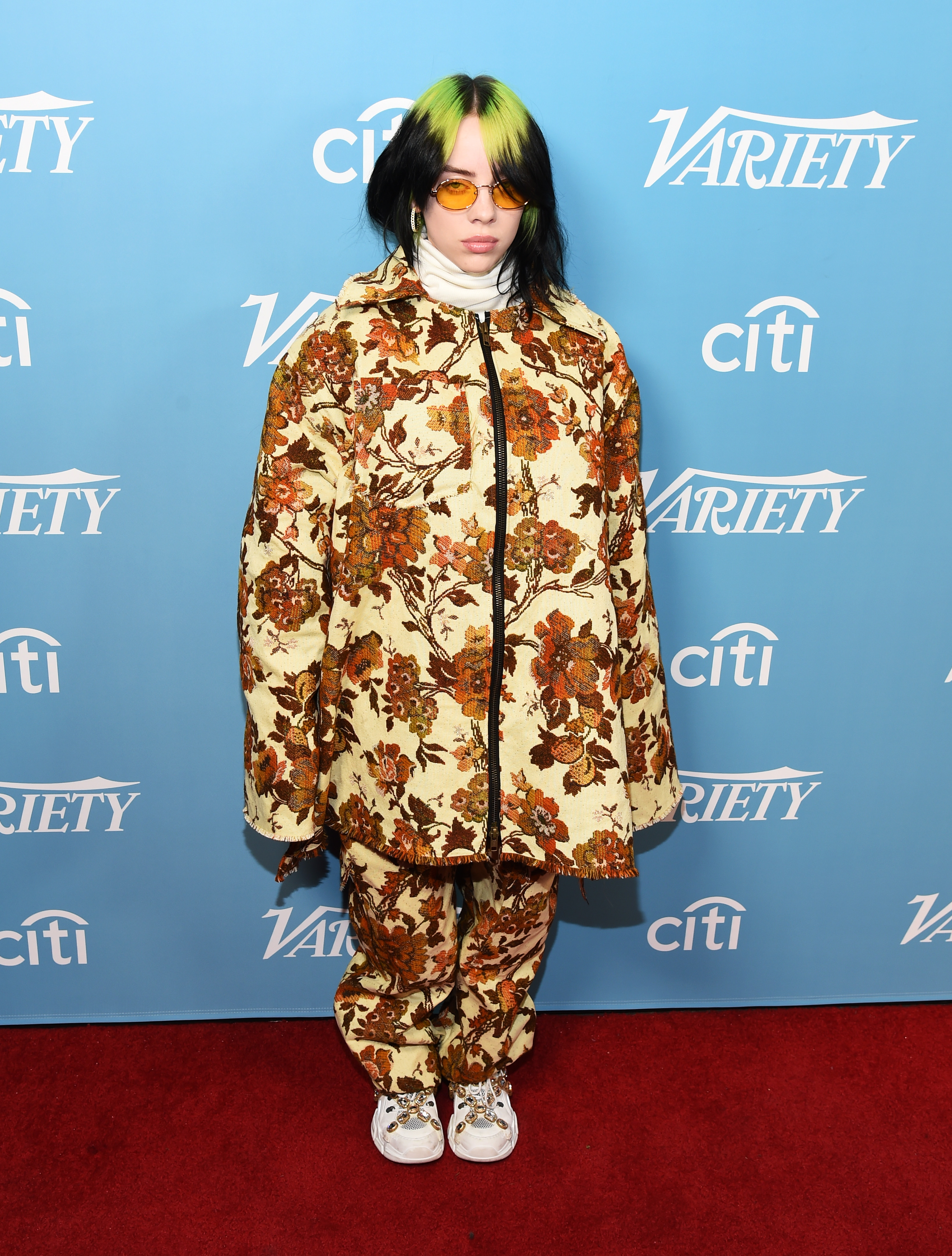 Billie Eilish no tapete vermelho. Ela usa um conjunto de estampa floral e tênis. Ela olha para a câmera e não sorri.