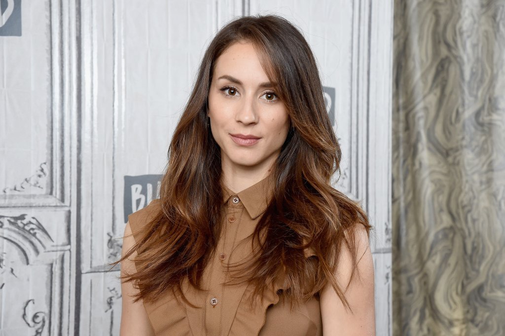 Troian Bellisario usando regata em tom de marrom com botões e cabelo solto; a atriz sorri levemente