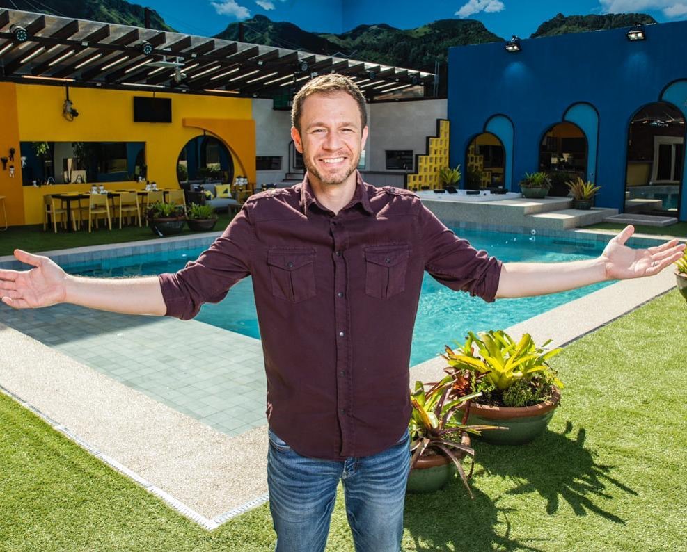 Tiago Leifert na casa do BBB20 com os braços abertos sorrindo na frente da piscina