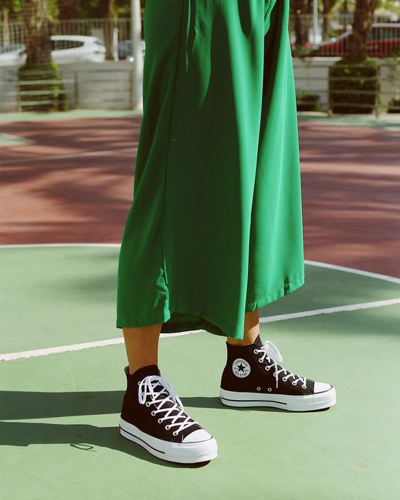 Montagem com foto em fundo de tintas em vermelho, rosa e verde com estrelinhas douradas em cima. Na foto, dá para ver os pés de uma garota usando tênis Chuck Taylor preto e branco da Converse e uma parte da saia midi verde dela.