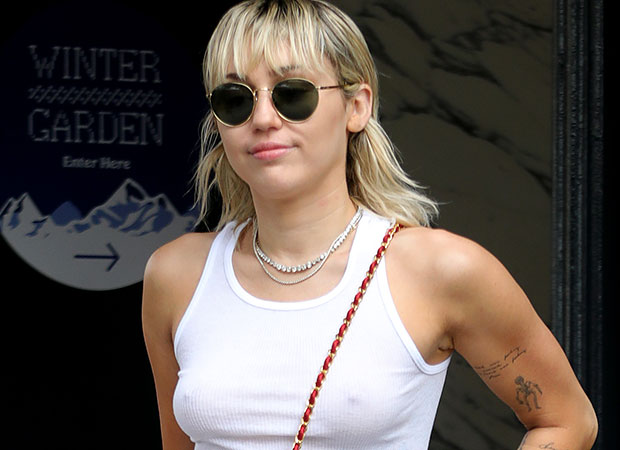 Miley cyrus com óculos escuro, regata branca, óculos preto, cabelo loiro e expressão sorridente.