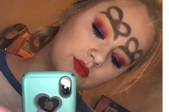 sobrancelha-de-laço Garota tirando foto com celular em frente ao espelho. Ela olha para baixo e sua maquiagem roxa, laranja e amarela dos olhos aparece. Ela tem um desenho de laço em cima de cada sobrancelha.