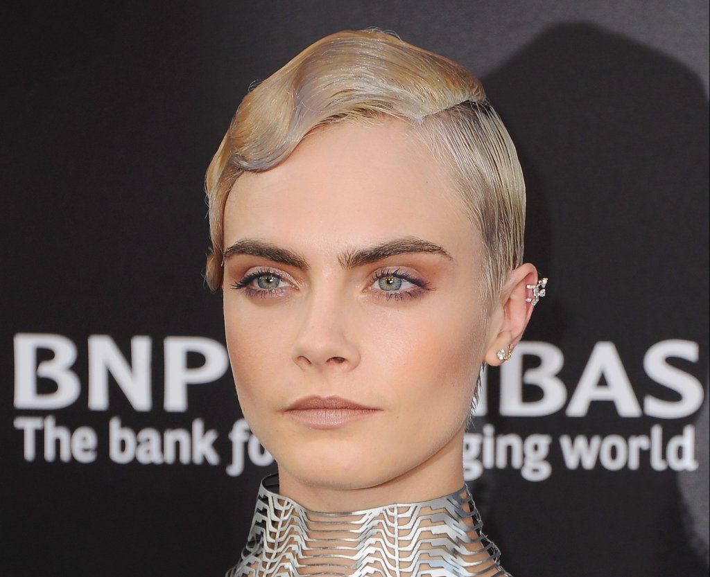 Cara Delevingne posando para foto com o cabelo penteado com gel; a expressão da modelo é séria e ela usa acessórios prateados