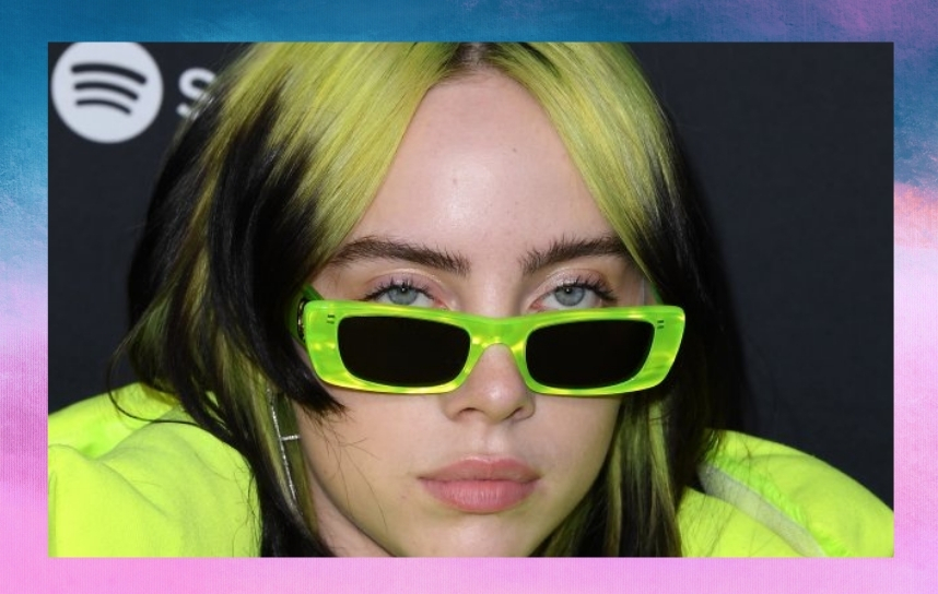 close no rosto de Billie Eilish. Ela está com expressão séria e usa jaqueta e óculos de sol com armação verde