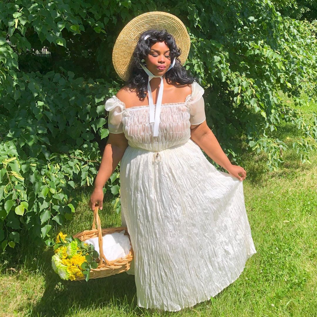 garota no campo usa vestido branco com decote ciganinha a saia rodada, além de um chapéu com uma fita amarrada no queixo