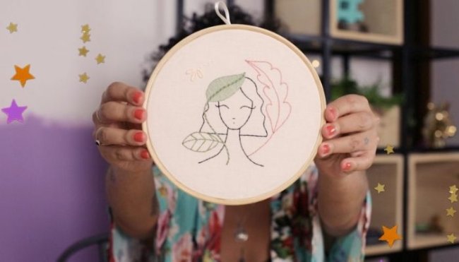 Bordado Mulher segura bastidor branco com desenho de um bordado de uma jovem com cabelo curto. Não conseguimos ver o rosto de quem segura o bordado, mas, conseguimos ver suas unhas pintadas de vermelho e sua camisa com detalhes florais.