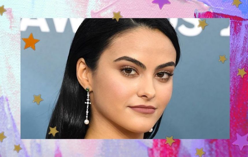 Foto com close no rosto da atriz Camila Mendes, que encara a câmera com uma expressão séria, ela usa brincos compridos com bolinha branca na parte final, batom com um tom marrom e suas sobrancelhas estão levemente arqueadas.