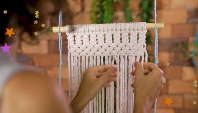 Na imagem só conseguimos ver duas mãos tecendo o macramê com linha branca em cima de um suporte e o fundo desfocado.