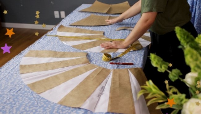 Molde de corte e costura Mesa com vários moldes de roupas, podemos ver mãos trabalhando nos moldes, fita métrica, canetas e lápis espalhados pela mesa.