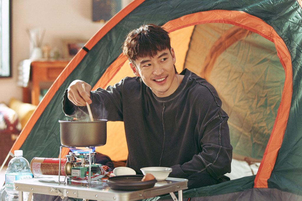 Cena de A Caminho do Céu; personagem está sorrindo e mexendo uma panela sentado na entrada de uma barraca de acampar