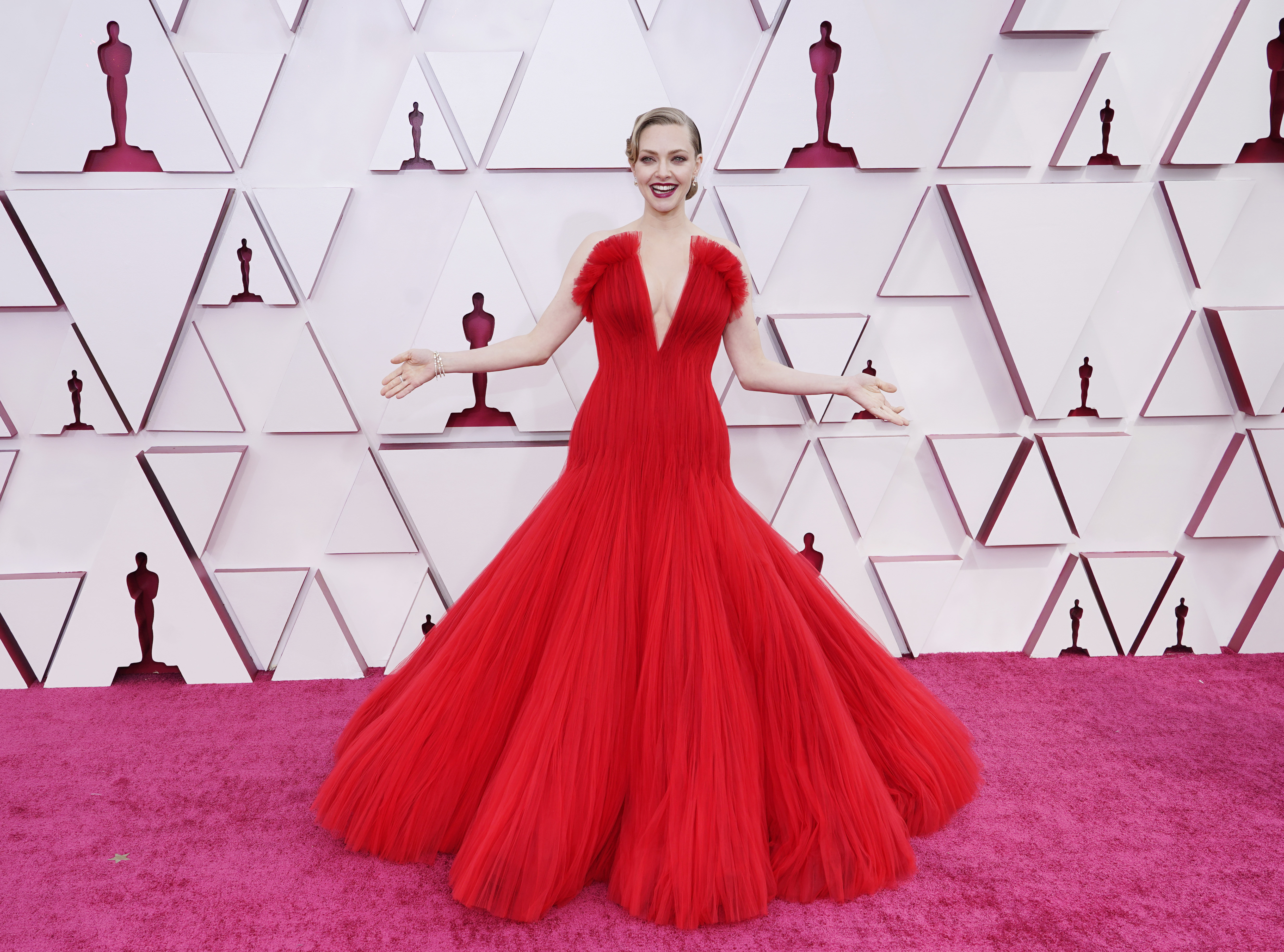 amanda-seyfried-oscar Amanda Seyfried no Oscar 2021. Ela está usando um vestido vermelho de tule com saia volumosa e decote V profundo. Com batom vermelho e cabelo preso em um coque baixo clássico, ela está em pé, com os braços abertos e sorrindo.