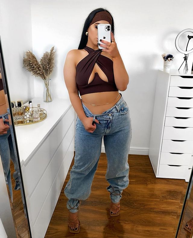 Mulher usando top cropped marrom com recorte no colo, calça jeans baggy e sandália marrom de tiras com bico quadrado. Ela também usa um cinto fino dourado na barriga, está em pé em frente ao espelho, com o celular na frente do rosto, e uma mão segurando no passador da calça.