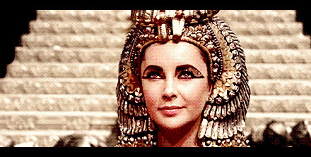Cleopatra Gif da atriz Elizabeth Taylor como Cleopatra usando adornos dourado ao redor da cabeça delineado grosso com detalhes dourados ao redor dos olhos e piscando para a câmera.