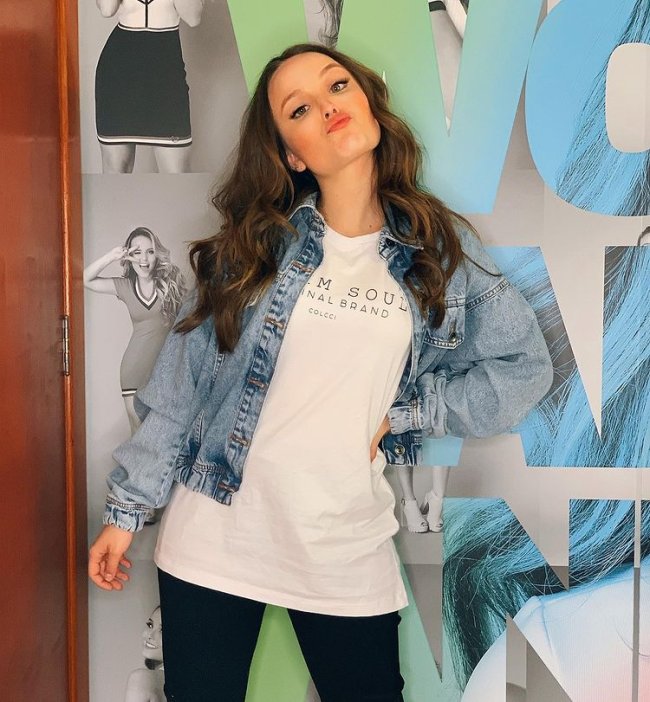 Larissa Manoela fazendo biquinho para foto, usando camiseta branca, jaqueta jeans e calça legging preta com uma das mãos apoiada na cintura