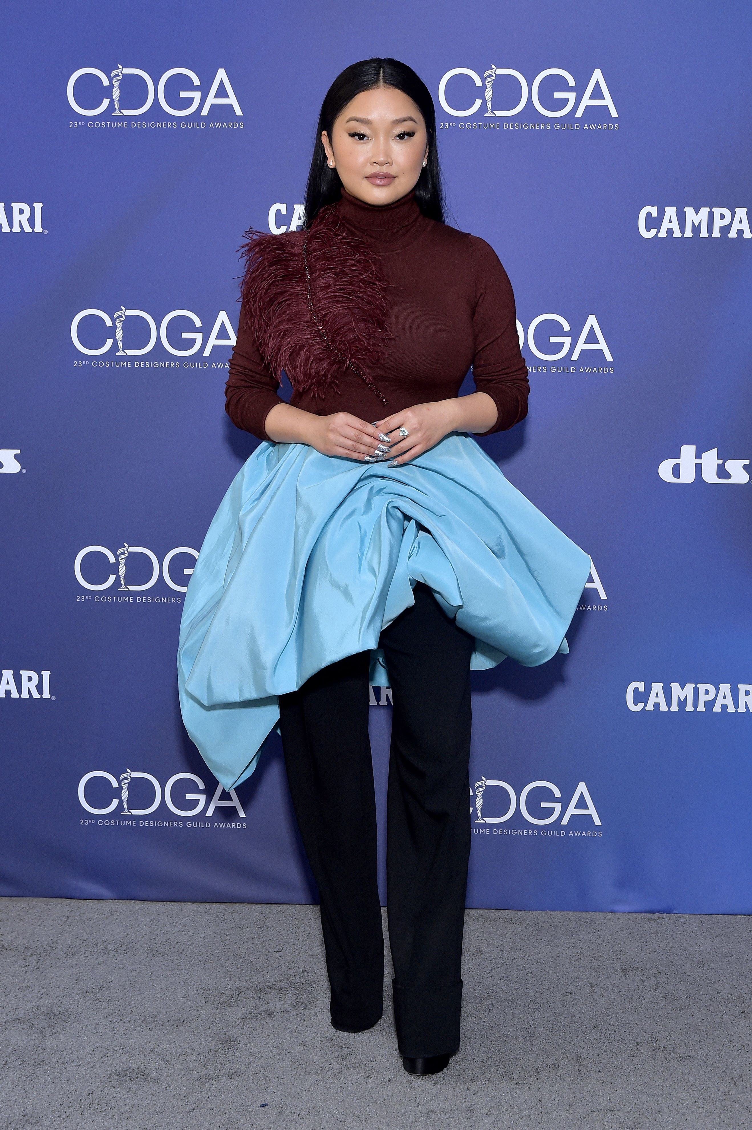 23rd Costume Designer Guild Awards Lana Condor usando suéter bordô de gola alta que tem uma aplicação de plumas em um dos ombros, uma calça pantalona azul-marinho e, por cima, uma saia azul turquesa volumosa e assimétrica. Ela está olhando para frente, com a expressão séria, os braços dobrados e as mãos unidas na frente do corpo.