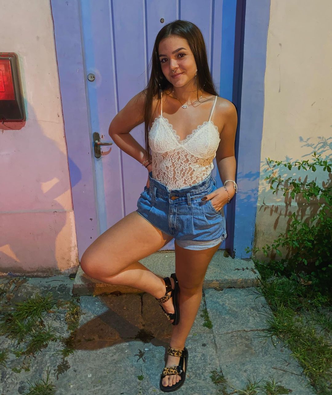 look-mel-maia-jeans-05 Mel Maia usando look com body de renda branco, short jeans modelo clochard e uma papete preta com estampa de oncinha nos pés. Ela posa em pé, com uma das pernas dobradas e apoiada na outra, formando um quatro, uma das mãos no bolso, e a outra na cintura. Ela está sorrindo, sem mostrar os dentes.