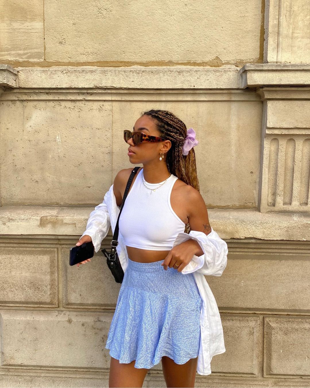 look-top-cropped-branco-amaka.hamelijnck- Garota usando top cropped branco com saia rodada azul e camisa branca por cima. Ela está com uma bolsa preta pendurada em um dos ombros, segurando o celular em uma das mãos, cabelo preso em um rabo com scrunchie lilás e óculos de sol. Ela está olhando para o lado.