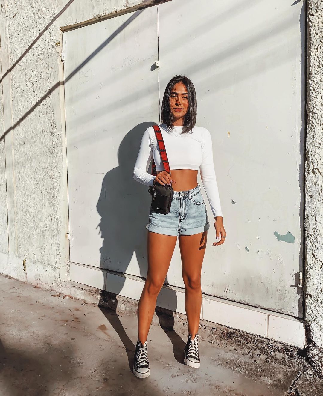 look-top-cropped-branco-brendapabreu Garota usando top cropped branco de manga longa com short jeans e tênis All Star preto e branco de cano alto. Em pé, ela está segurando uma minibolsa de alça longa em um dos ombros.