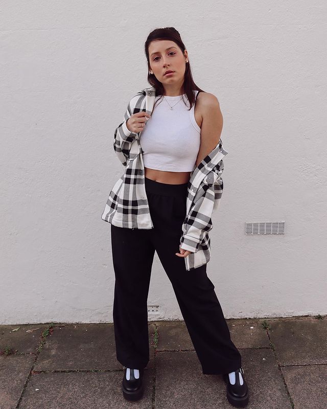 look-top-cropped-branco-brunaqbarros Garota usando top cropped branco com calça preta de cintura alta, sapato estilo boneca preto e meia branca. Por cima, ela usa um casaco xadrez preto e branco. Está em pé, com expressão facial séria, e uma das mãos segurando o casaco.