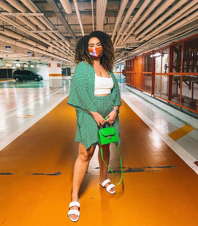 Garota usando top cropped branco com kimono e bermuda estampados em verde com bolinhas brancas. Ela está em pé, usa uma rasteirinha branca de duas tiras, segura uma bolsa verde neon na frente do corpo e está com uma máscara de proteção laranja e rosa.