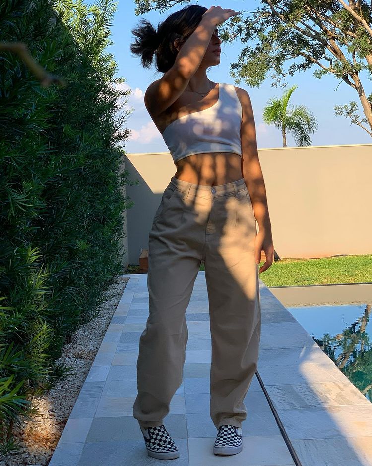 look-top-cropped-branco-gabitrassi Garota usando top cropped branco com alça de um ombro só com uma calça baggy bege e tênis xadrez preto e branco. Ela está em pé, com a cabeça virada para o lado e uma das mãos na frente do seu rosto.