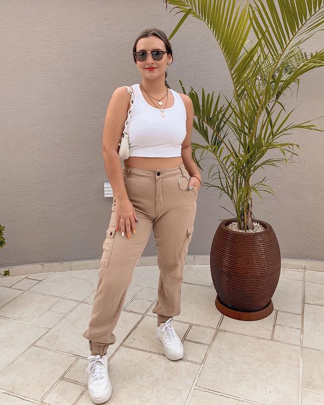 look-top-cropped-branco-gihbertozzi Garota usando top cropped branco com calça cargo bege e tênis branco. Ela está em pé, com uma bolsa branca pendurada em um dos ombros, óculos de sol e colares de correntes. Sua expressão facial tem um leve sorriso e uma das mãos está no bolso.
