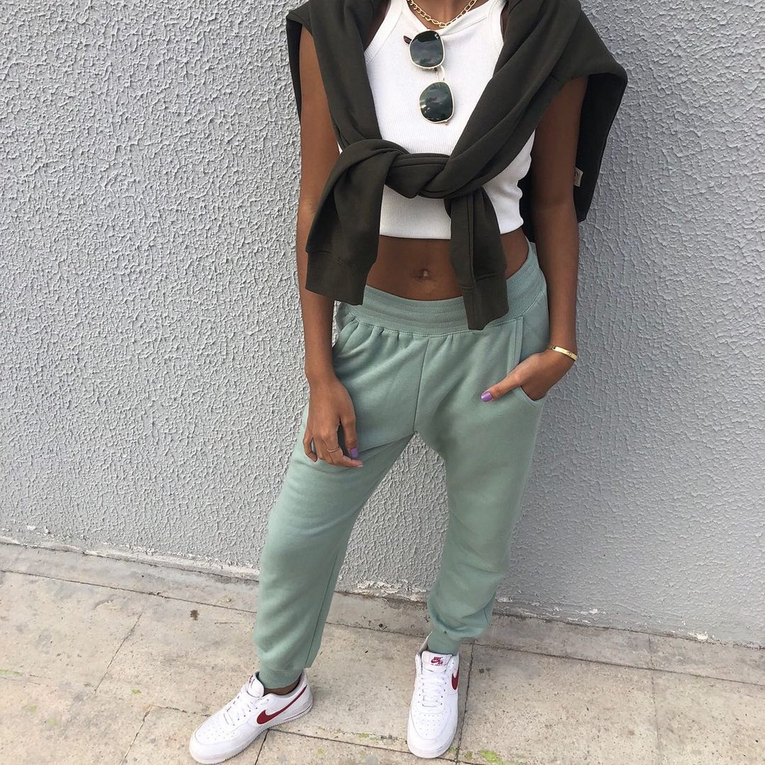 look-top-cropped-branco-julinha_sanches Garota usando top cropped branco com calça de moletom verde, tênis branco e suéter preto amarrado nos ombros. Tem um óculos de sol pendurado na gola da blusa, e a foto pega apenas do pescoço para baixo.