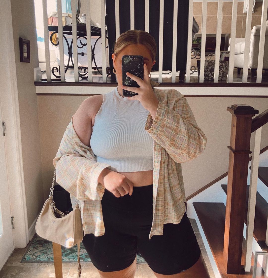 look-top-cropped-branco-lauren_wambolt Garota usando top cropped branco com bermuda ciclista e camisa xadrez laranja e cinza por cima. Um dos braços segura uma bolsa bege com alça de corrente, e a outra mão segura o celular para tirar foto em frente ao espelho.