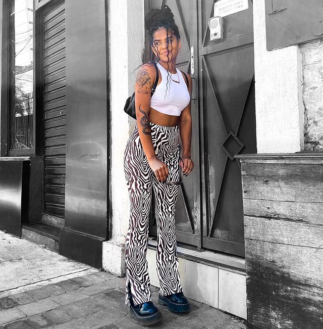 look-top-cropped-branco-soeirojuliana Garota usando top cropped branco com calça preta e branca de zebrinha, coturno preto e uma bolsa preta pendurada em um dos ombros. Ela está em pé, com as duas mãos na frente do corpo segurando um óculos de sol, enquanto olha para o lado.