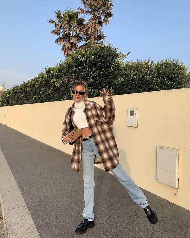 look-top-cropped-branco-sofiamcoelho- Garota usando top cropped branco com calça jeans, cinto preto, oxford preto e camisa xadrez marrom e branca por cima do look. Ela está com uma das pernas levantadas, uma mão segurando um pacote, a outra fazendo o símbolo de paz e amor com os dedos. Ela ainda usa um óculos de sol e sorri.