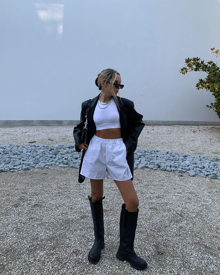 Garota usando top cropped branco com short branco de cintura alta, bota tratorada preta que vai até o joelho e um casaco de couro preto por cima. Ela está em pé, usando óculos de sol e uma bolsa preta em um dos ombros. Sua cabeça está inclinada para um dos lados, e um dos joelhos está dobrado.