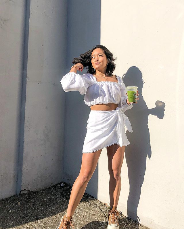look-top-cropped-branco-sophiesinds- Garota usando top cropped branco com mangas longas e bufantes e saia branca com amarração na lateral. Nos pés, um tênis marrom de cano alto. Com uma das mãos, ela segura um suco verde, enquanto a outra mexe no cabelo.