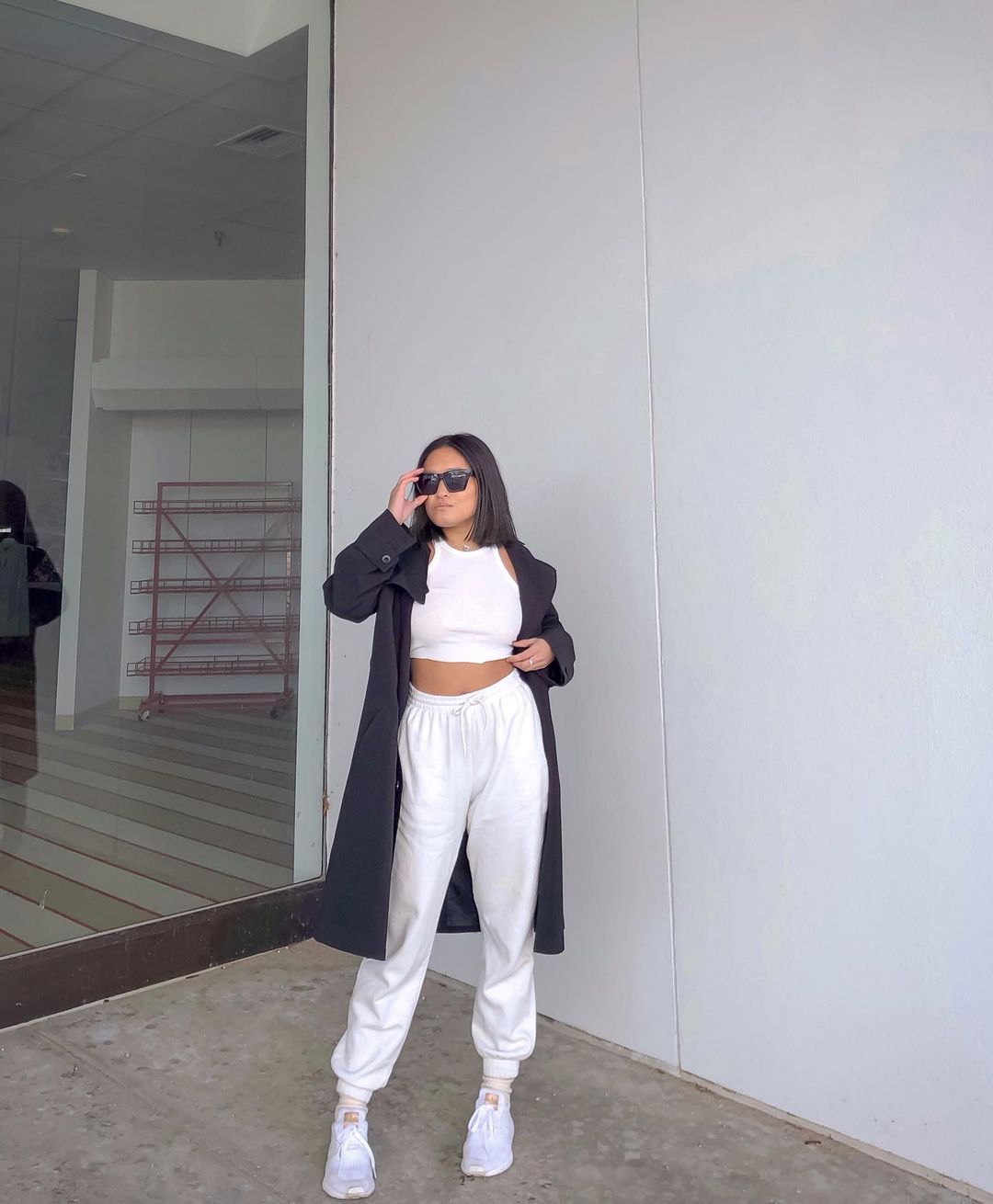 look-top-cropped-branco-sophiesinds Garota usando top cropped branco com calça de moletom e tênis brancos. Ela está com um sobretudo preto por cima do look, e uma das mãos segura os óculos escuros, enquanto a outra está na cintura.