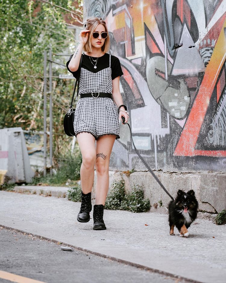 Garota usando look com macaquinho xadrez vichy preto e branco e camiseta preta de manga curta por baixo, além de um cinto preto. Nos pés, um coturno preto, uma bolsa preta pendurada em um dos ombros, e usando óculos de sol. Ela está em pé, com uma das mãos no cabelo, e a outra segura uma coleira de um cachorro pequeno de pelos em preto e branco.