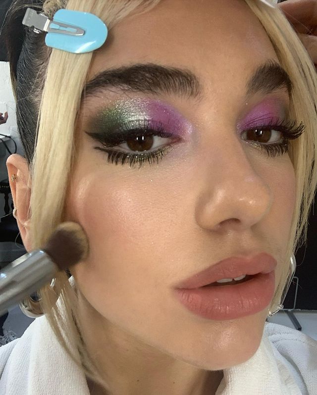 maquiagem-dua-lipa-01 Dua Lipa usando maquiagem colorida com sombras verde, prata e roxa, além de um delineado preto gatinho. A foto mostra apenas o rosto da cantora. Ela está com a boca levemente aberta, expressão séria e um pincel toca uma de suas bochechas.