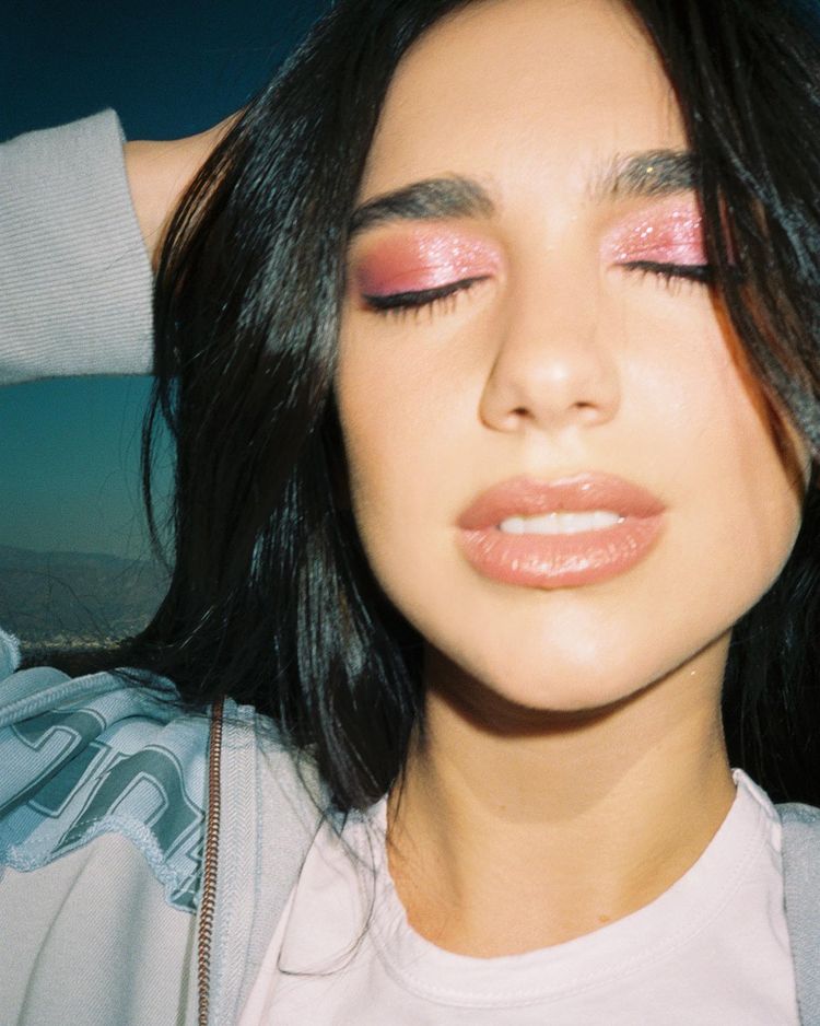 Dua Lipa usando maquiagem com sombra rosa de brilhos e gloss marrom. Ela está com os olhos fechados, boca levemente aberta e uma das mãos na cabeça.