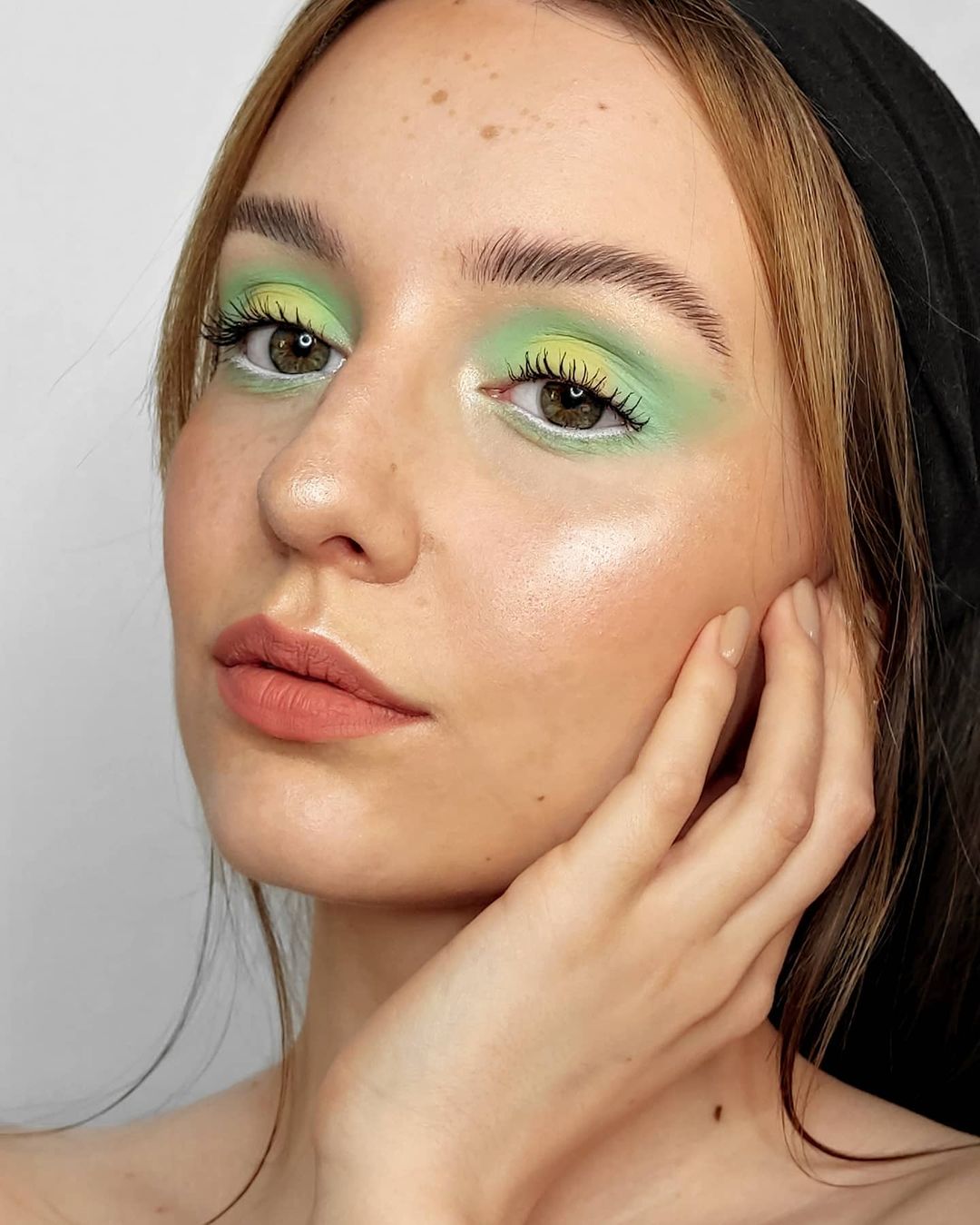 maquiagem-olho-verde-a.niaszlaga Garota usando maquiagem com diferentes tons de verde. Na foto, só dá para ver seu rosto e sua mão, apoiada no rosto. Ela está com a cabeça levemente voltada para o lado, expressão facial séria e olhando para o lado.