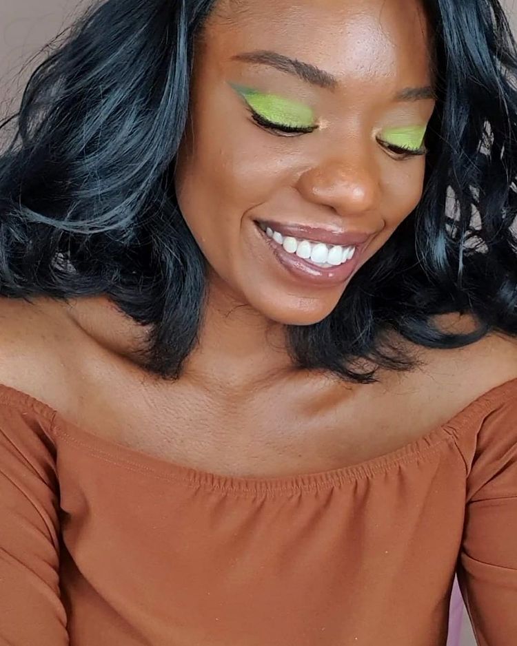 maquiagem-olho-verde-damatamakeup Garota usando sombra verde nos olhos e blusa marrom ombro a ombro. Ela está olhando para baixo e sorrindo. Seu cabelo é preto ondulado.