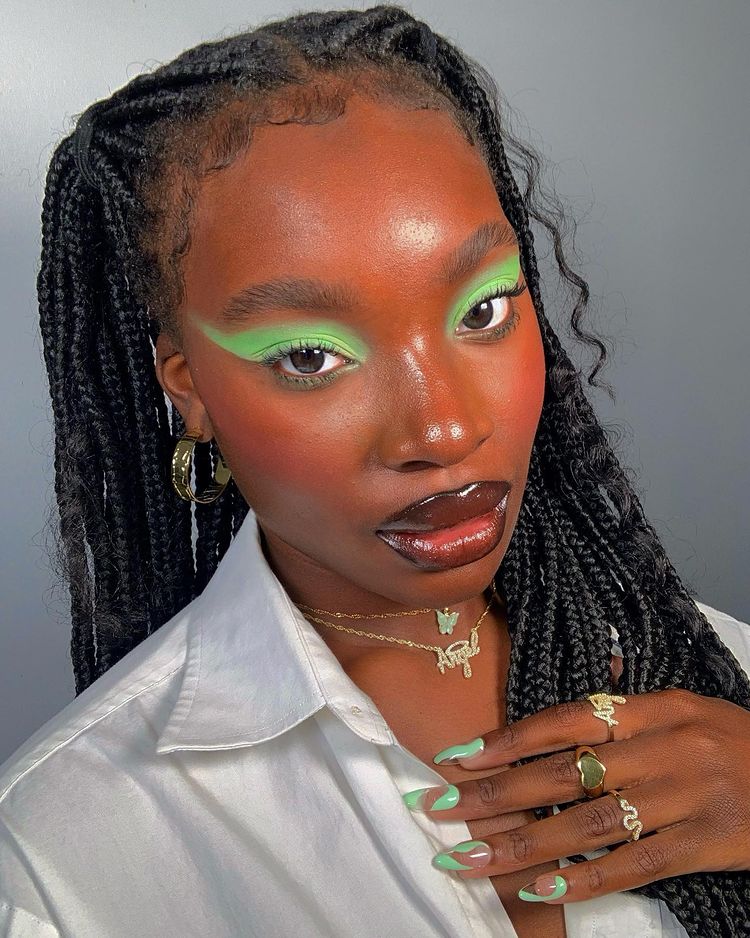 maquiagem-olho-verde-isabelleikpeme Garota usando maquiagem com sombra verde, uma camisa branca e mix de colares dourados. Sua mão, com as unhas com nail art verde, está apoiada perto do pescoço. Sua expressão facial é séria.