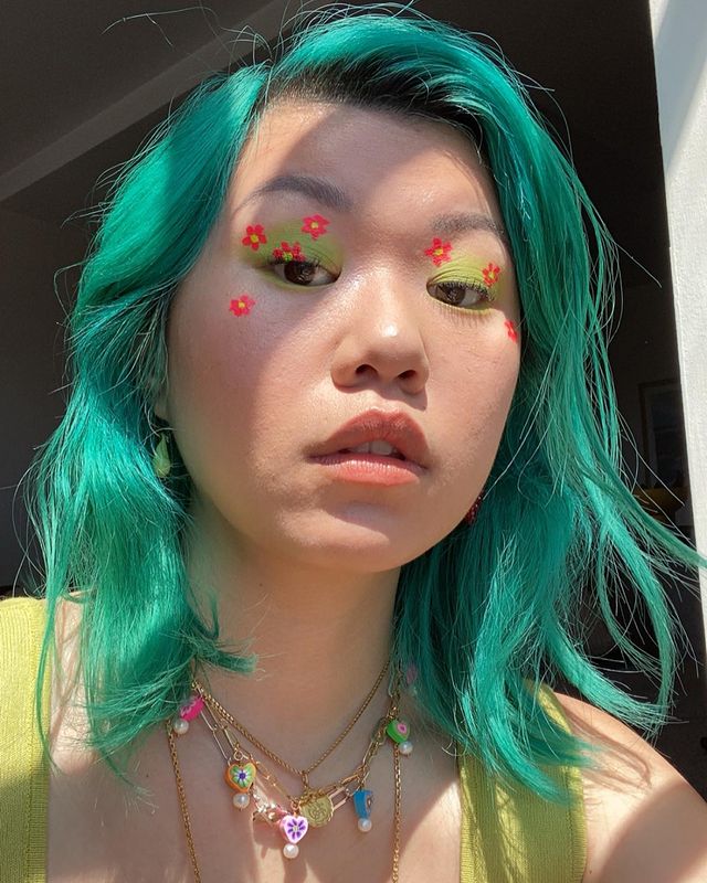 maquiagem-olho-verde-mianne.chan Garota usando maquiagem com sombra verde e florzinhas laranjas. Seu cabelo e sua regata são verdes e ela usa mix de colares. Ela está olhando para baixo, com expressão facial séria e boca levemente aberta.