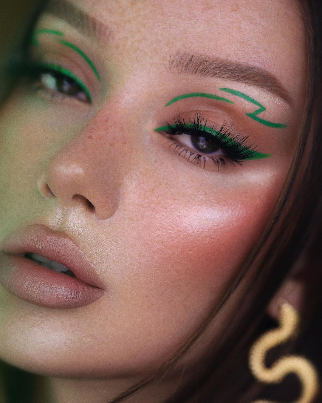 maquiagem-olho-verde-olgadann Garota usando maquiagem com delineado gatinho verde e um delineado gráfico na parte acima da pálpebra. Na foto, só dá para ver o rosto, que tem uma pele iluminada, blush bem mercado e batom cor de boca. A expressão facial é séria, com a boca levemente aberta.