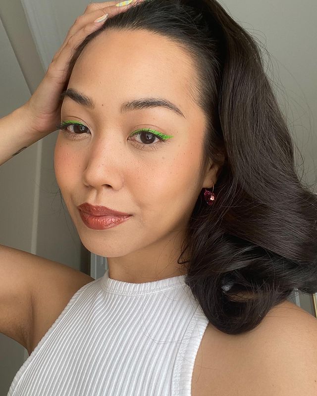 maquiagem-olho-verde-overglowedit Garota usando blusa branca e delineado verde nos olhos. Uma das mãos está apoiada na cabeça e sua expressão facial é séria. Seu cabelo está preso em um rabo e na foto só dá para ver do ombro para cima.