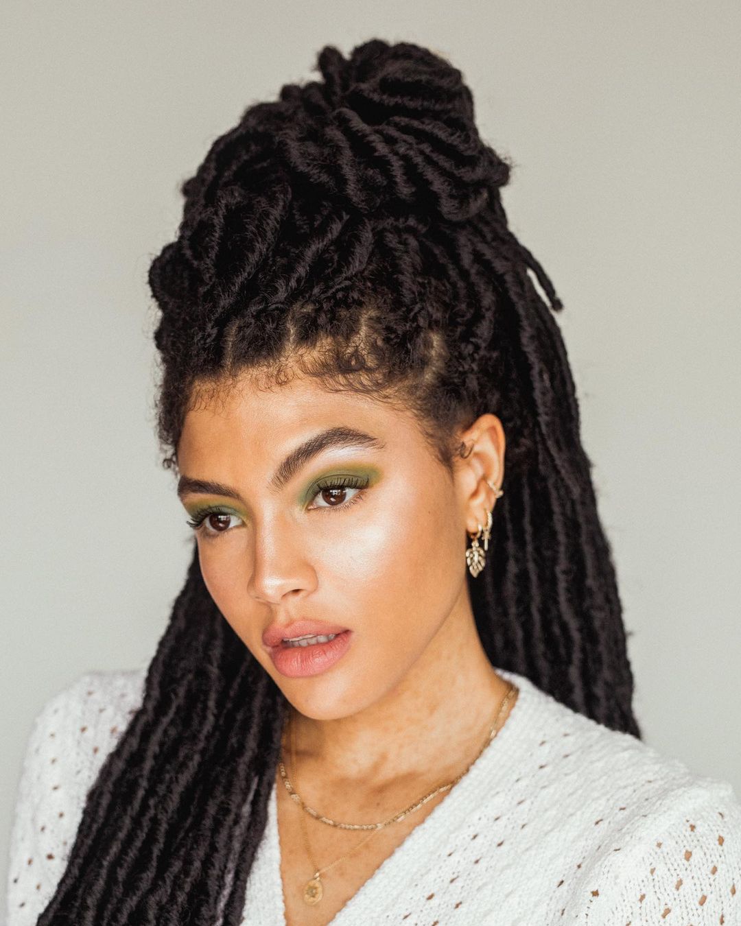 maquiagem-olho-verde-samiorenelda Garota usando maquiagem com sombra verde oliva e suéter branco. Ela está olhando para baixo, com expressão facial séria e a boca levemente aberta. Seu cabelo está com box braids, e a foto só mostra dos ombros para cima.